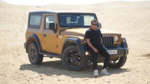Home 6 A man leans on a Mahindra Thar SUV in a desert setting under bright sunlight.